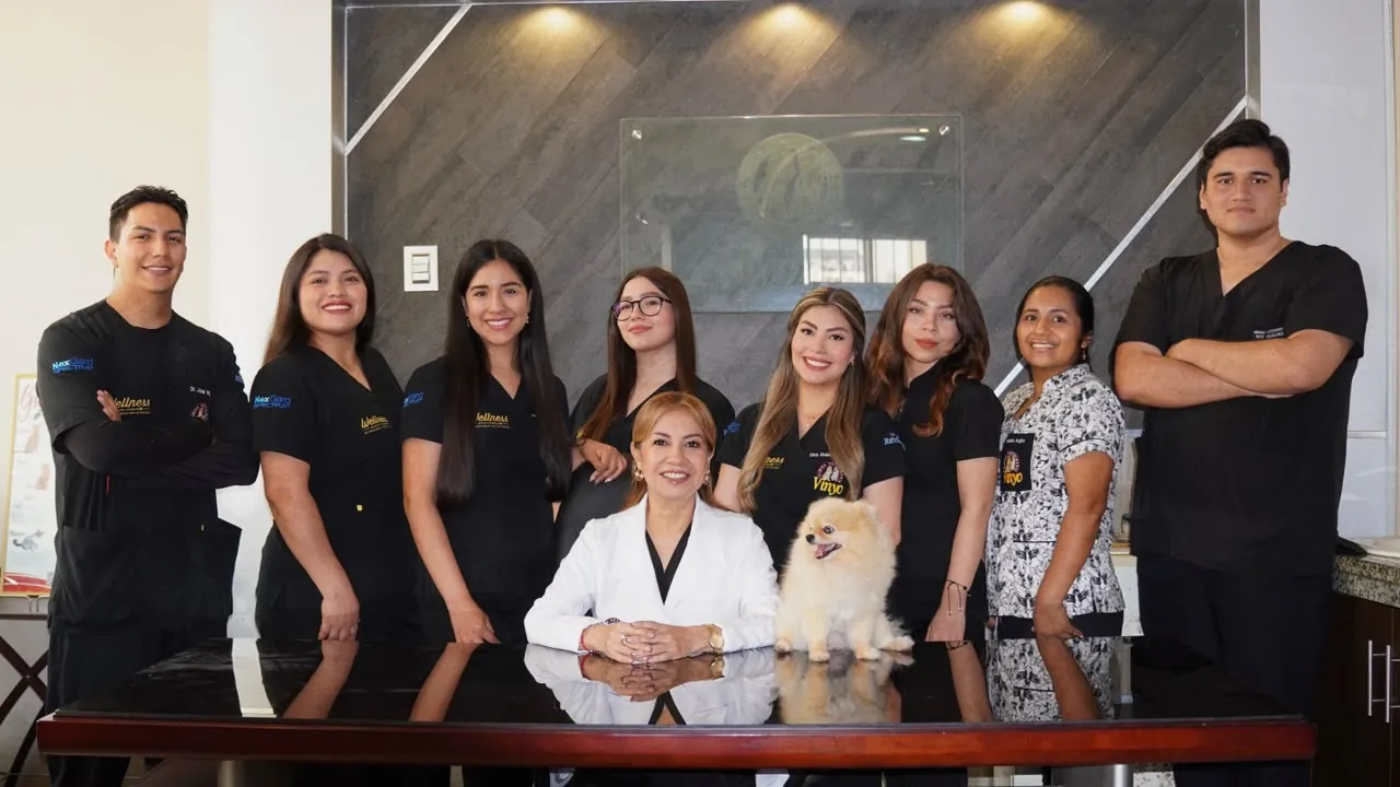 El equipo de la clínica, con uniforme negro, de pie detrás de una mesa; al centro, una doctora con bata blanca sentada y un pomerania sobre la mesa